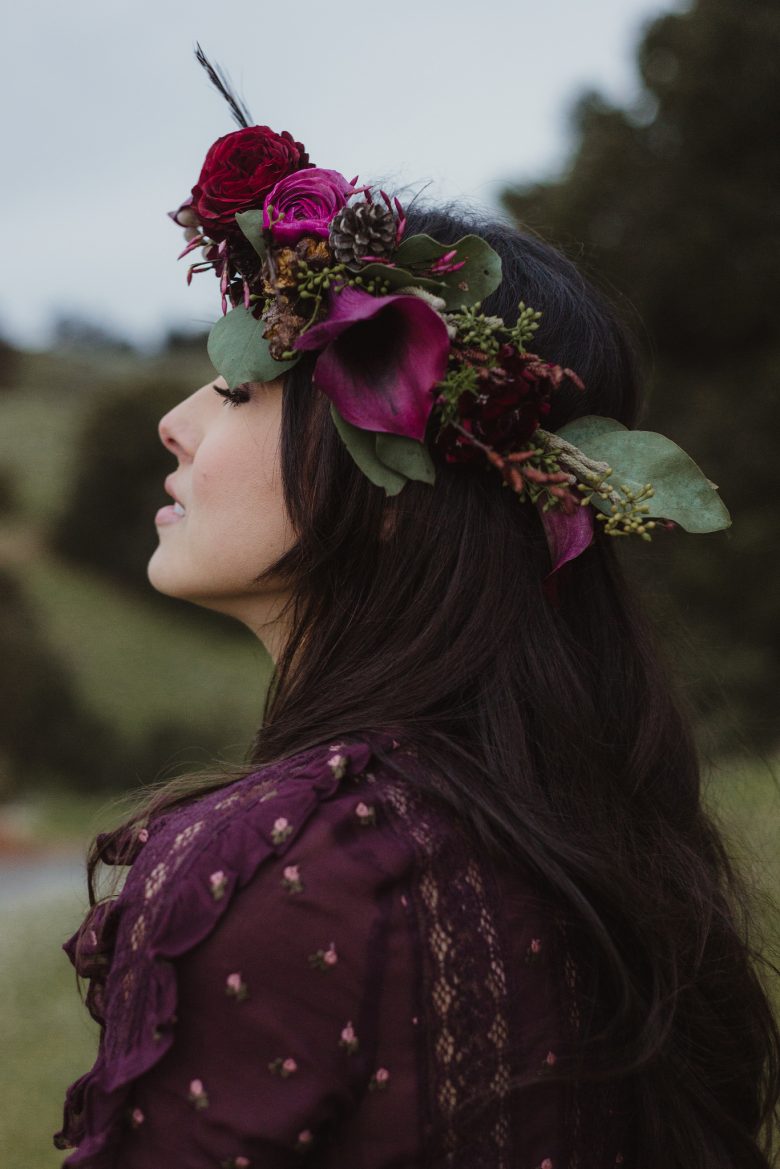 A raven haired bride stands thoughtfully with her eyes closed, wearing a flower crown that perfectly matches the her maroon wedding dress