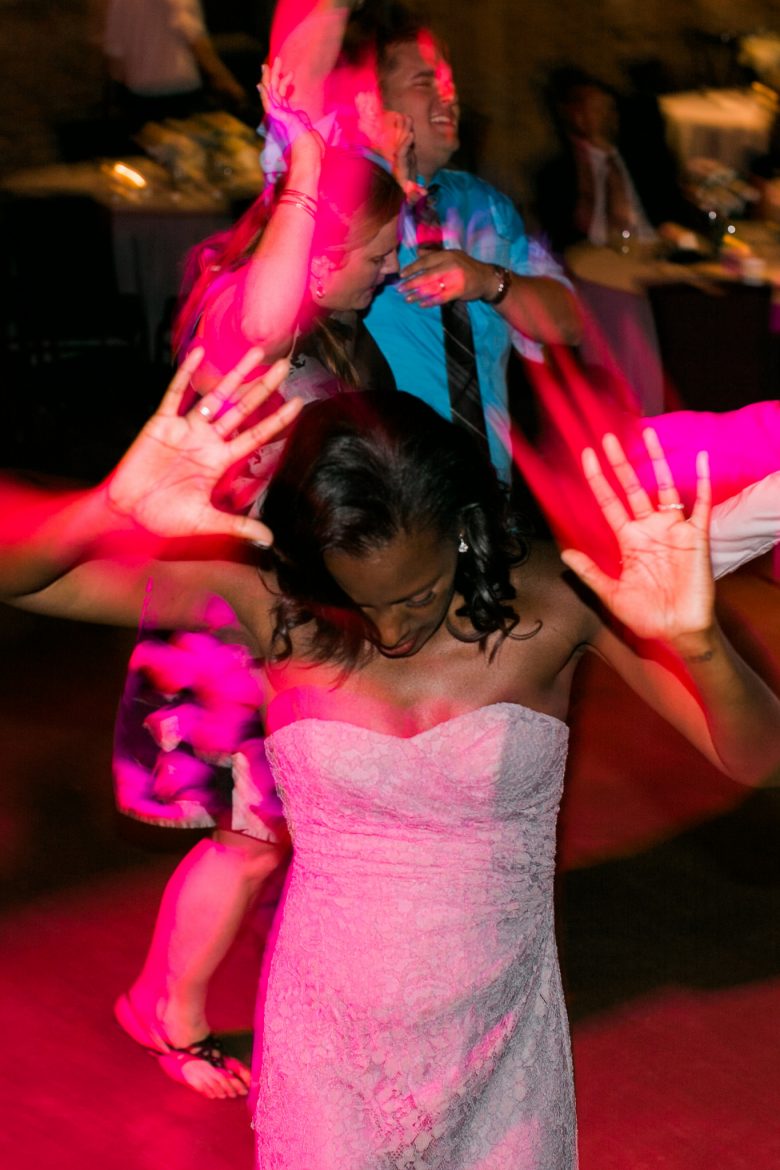A bride bathed in neon pink light on the dance floor, dances with her face down and hands up mid-motion