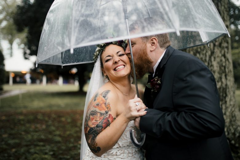 Groom kissing the cheek of smiling bride with colorful tattoo underneath clear umbrella near a tree