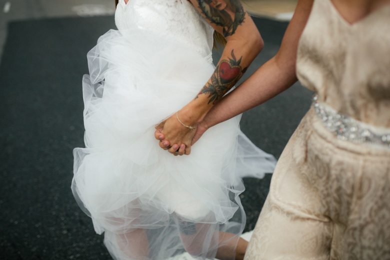 Two brides, one with tattoos on her arm, holding hands