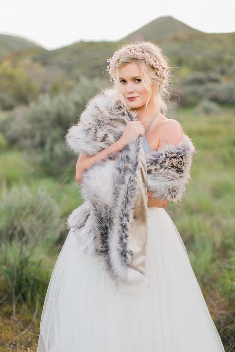 A bride stands in a grassy field wrapped in a luxurious faux fur shawl