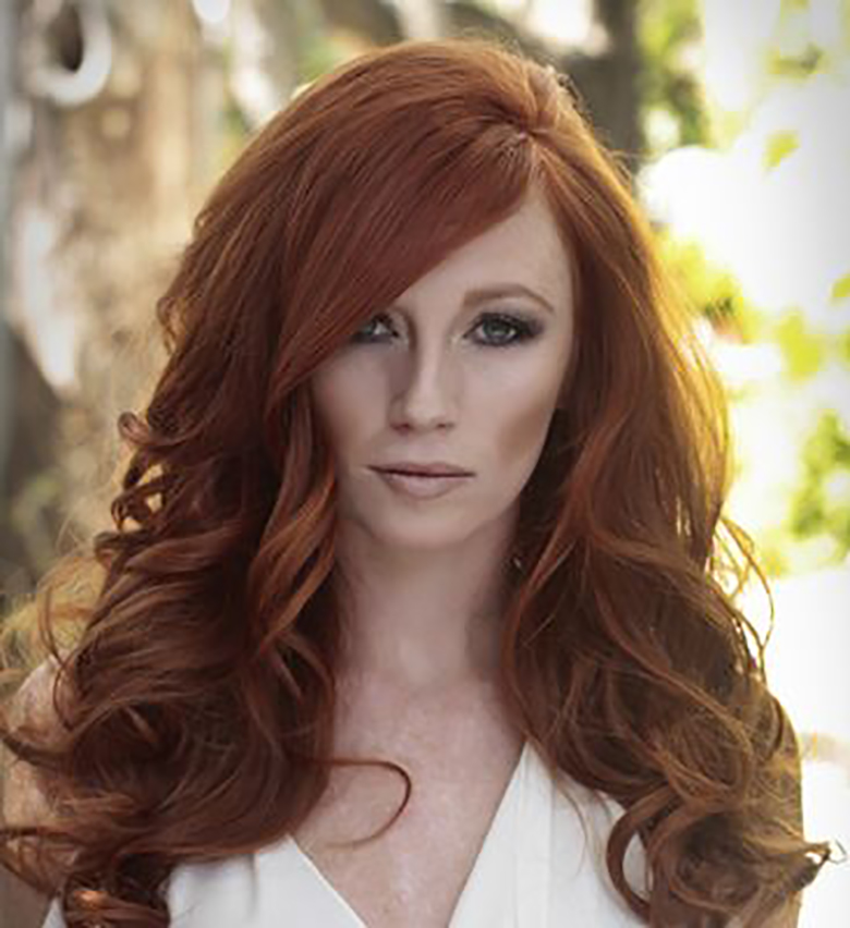 Woman with red hair with large, voluminous curls and a dramatic side-part bang with dramatic contour makeup and nude lip