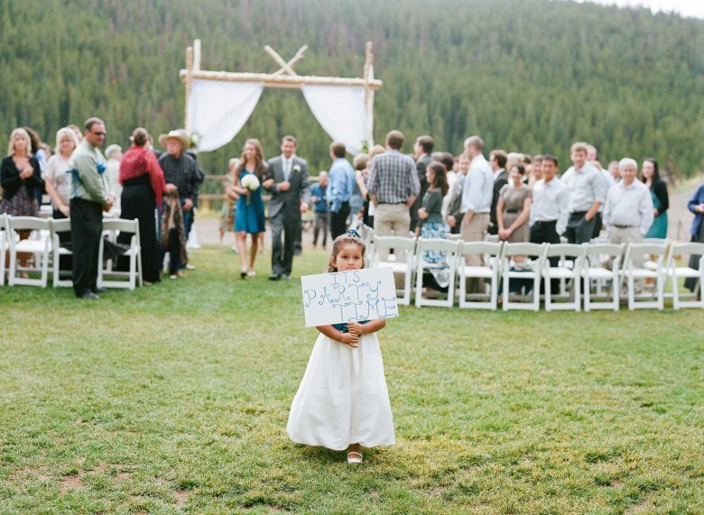 Young girl in white dress carrying a sign that reads It's Party Time down the aisle at an outdoor wedding