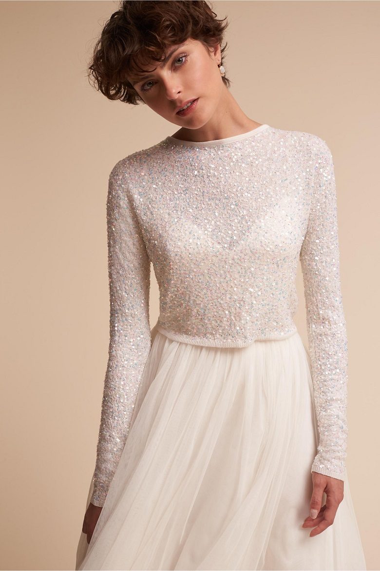 A bride stands with a while skirt and a white semi-sparkly long sleeve sheer sweater