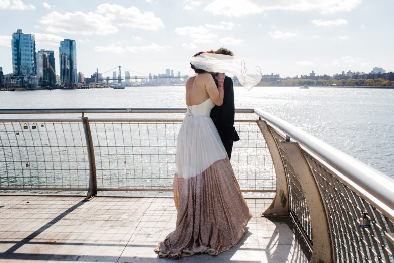 Bride and groom standing near water, overlooking skyline, with veil blowing in the wind, obscuring faces