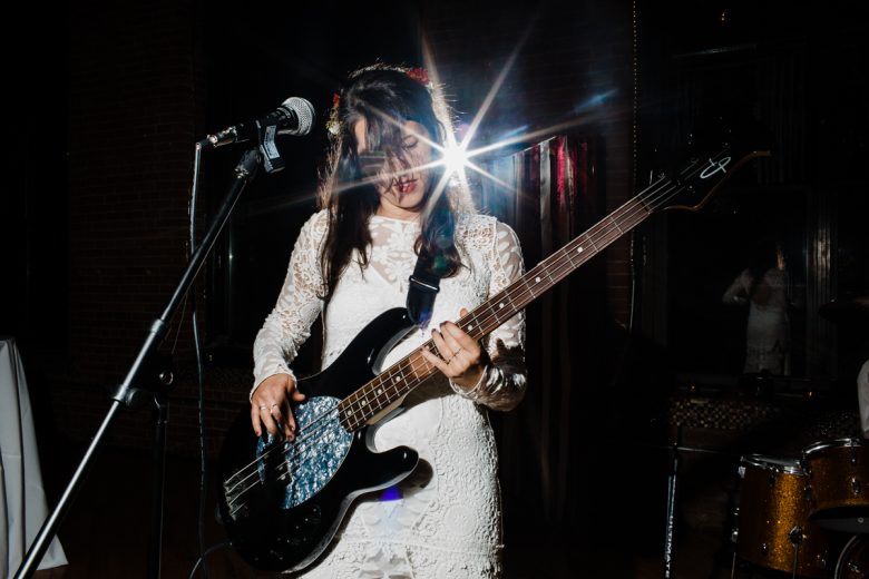 bride with long dark hair playing bass near microphone with light shining behind her