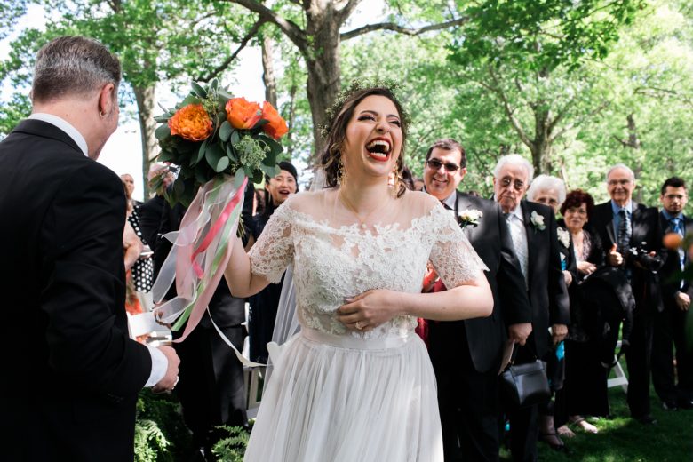 bride laughing holding a bouquet of orange flowers in front of besuited guests