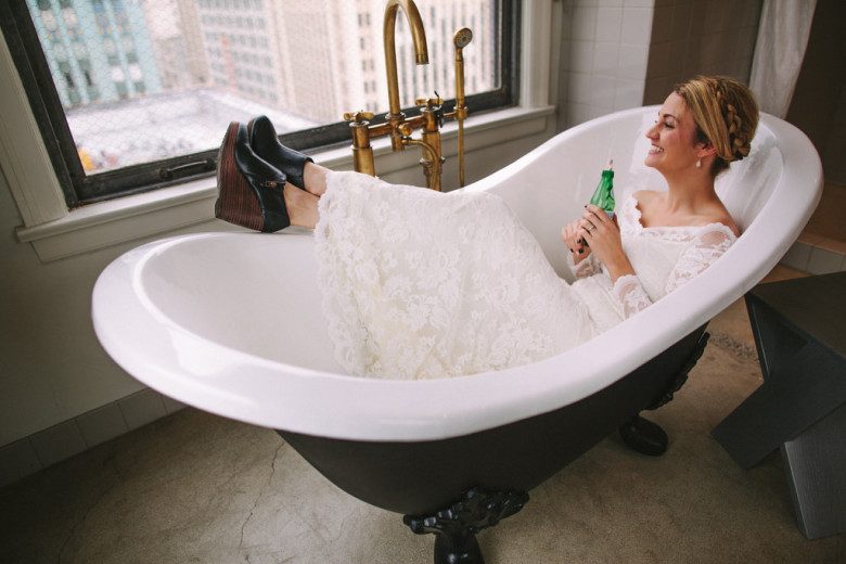 a bride sits in a claw foot tub while sipping from a green glass bottle and looking out at the city skyline
