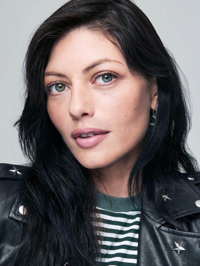 A raven haired woman wearing a leather jacket glances at the camera