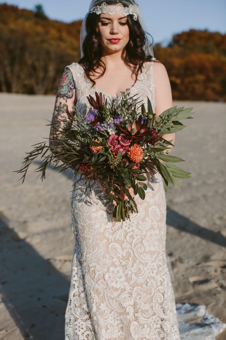 A bride stands on a beach, pensively looking at her bouquet.