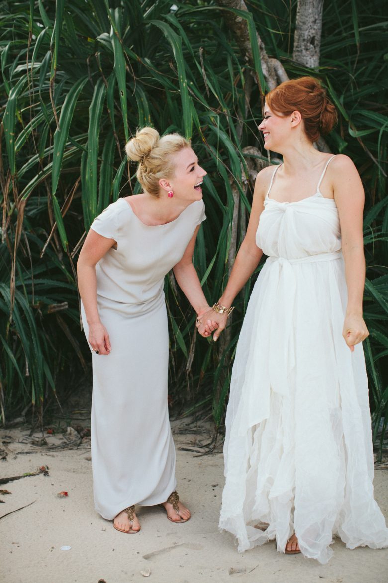 Two brides holding hands and laughing in front of tropical green plants