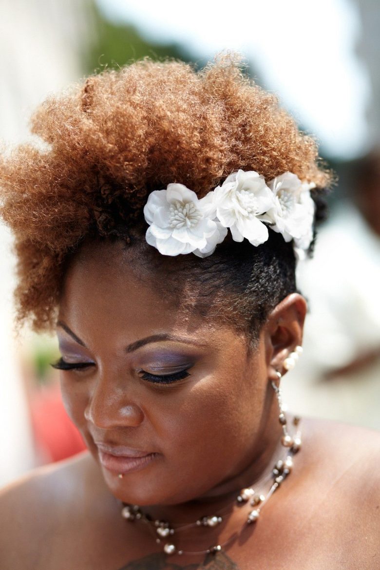 woman with tightly coiled natural hair, smooth on one side, piled on top and side, with white floral hairpiece 