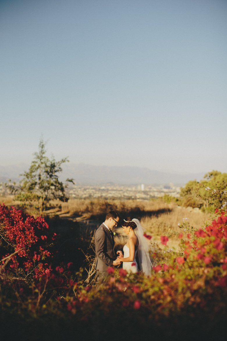 A wedding couple stand alone in a field facing each other and holding hands as the setting sun bathes them in light