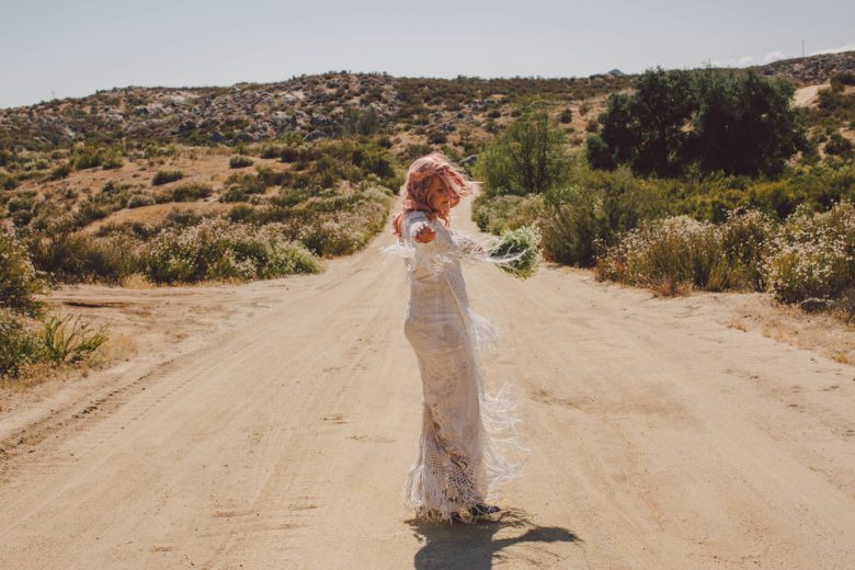 A bride stands in the middle of a dirt road, arms outstretched as the wind blows at the fringe on her dress 