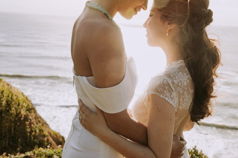 Two women in wedding dresses stand at the ocean, bathed in light from the setting sun they embrace each other.