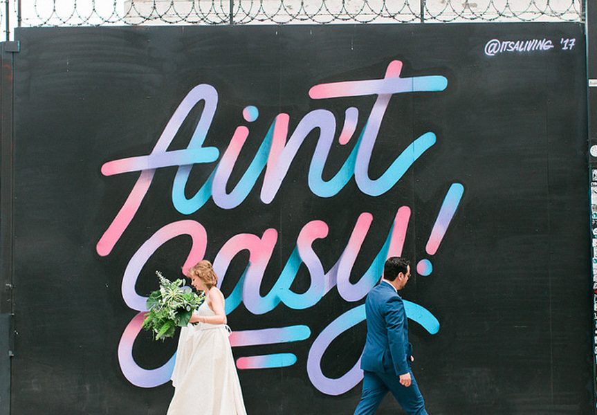 A couple walks away from each other in front of a wall with the words Ain't Easy painted on it