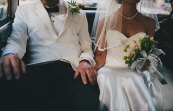 A wedding couple sit in the back seat of a car, holding hands.