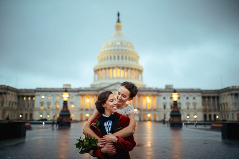 woman in red tux jacket being hugged by woman in wedding gown in front of the capitol building