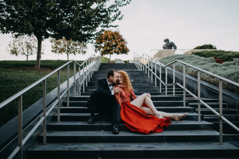 woman with red hair wearing a red gown sitting on steps leaning against man in black tux sitting beside her