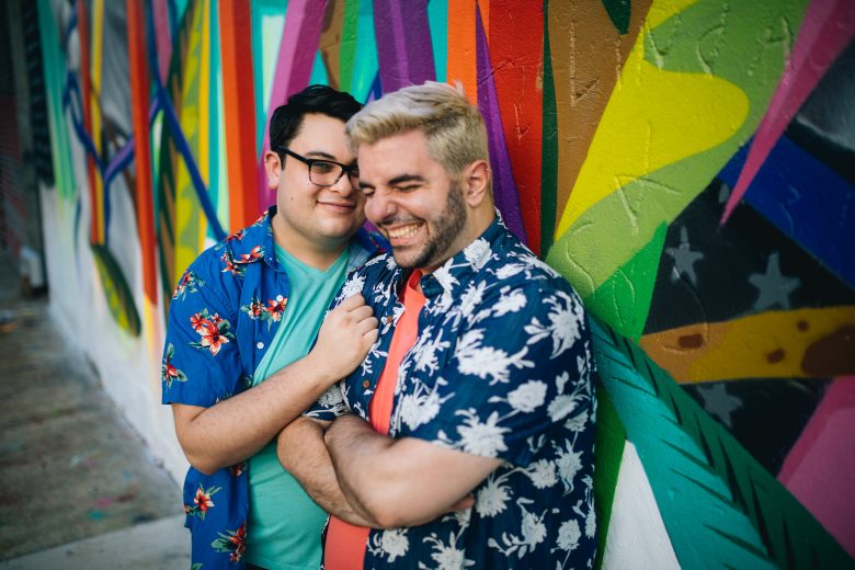 Two men laughing and embracing in front of a brightly colored wall