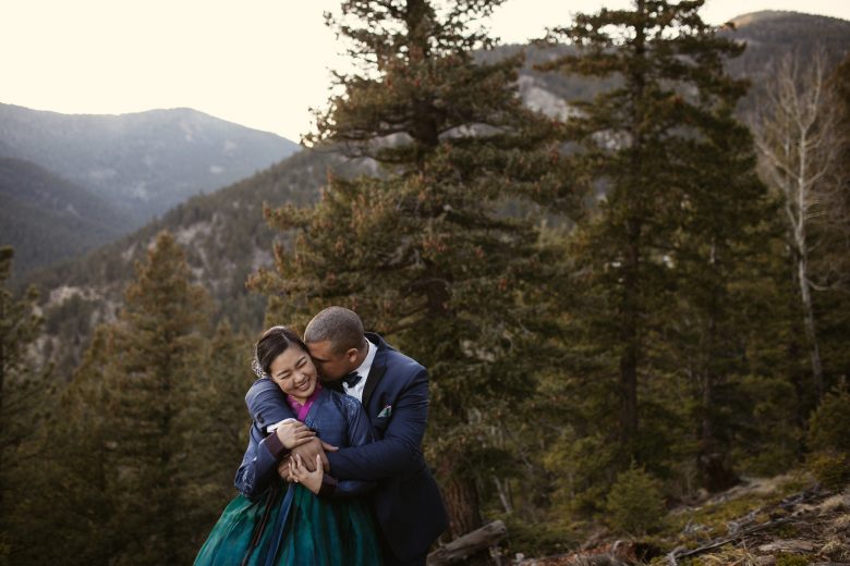 Man in tux kissing woman in blue, pink, and green gown in the forrest on a mountain
