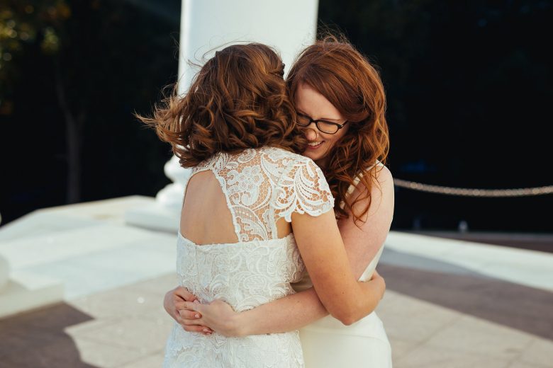 two brides hugging and smiling in front of a white pillar