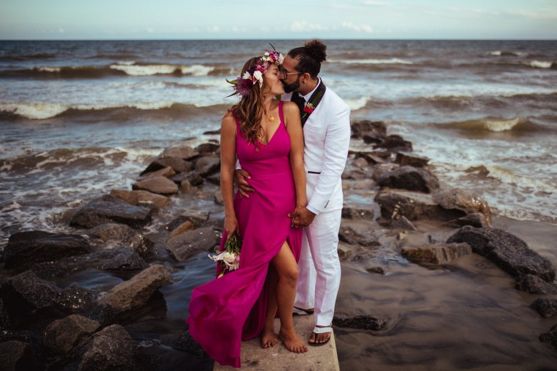 Woman with flower crown in bright pink dress and man in a white tux kissing on a rock on the beach in front of the ocean
