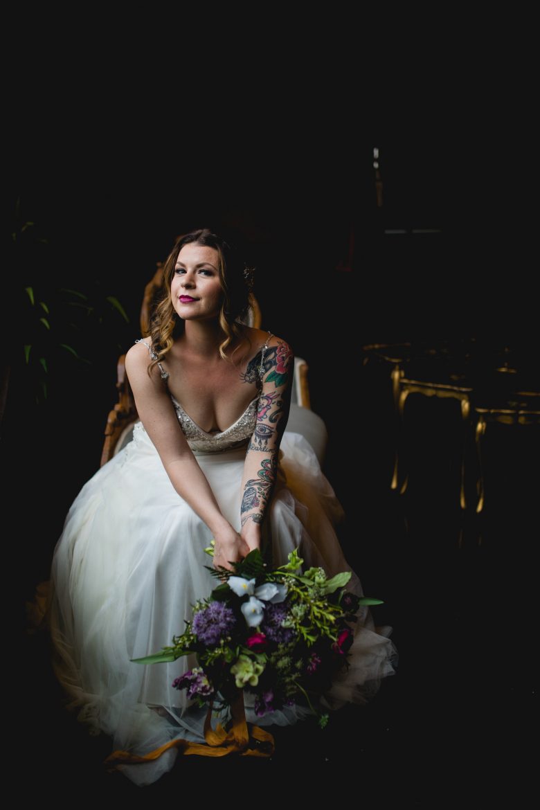 woman in wedding gown and sleeve of tattoos holding large colorful bouquet leaning forward in a chair in a dark room