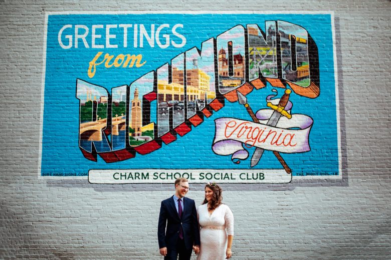 bride and groom holding hands and smiling in front of a wall that reads "Greetings from Richmond Virginia Charm School Social Club"