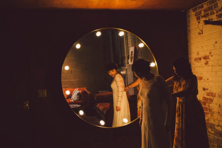 A bride stands beside a mirror as her wedding dress is zipped up from behind