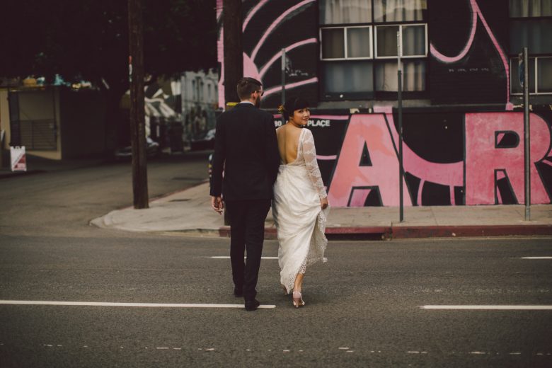 the wedding couple cross a city street as she looks over her shoulder back at the camera
