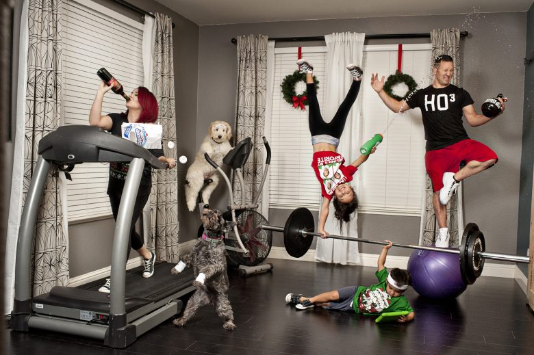 family portrait with woman on treadmill drinking wine from the bottle and eating powdered sugar mini donuts, which are falling out of the bag to an awaiting grey dog; child on the ground viewing tablet holds large barbell overhead, another child does a one-handed handstand on the barbell and shoots man with water from water bottle; man balances in tree pose on a yoga ball while holding an open beer growler, turning his head to avoid the squirt from the water bottle; white furry dog sits on a fan treadmill in the corner