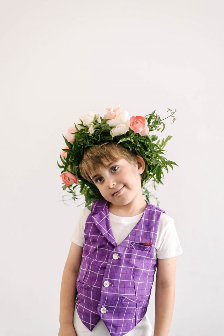 a young boy smirks while wearing a purple plaid vest and a flower crown