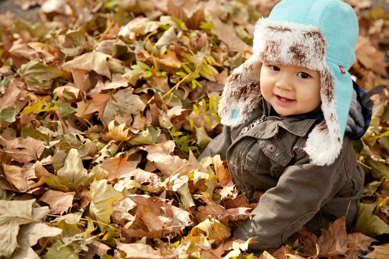 young child in a blue and fur flap cap sits in a pile of leaves, smiling