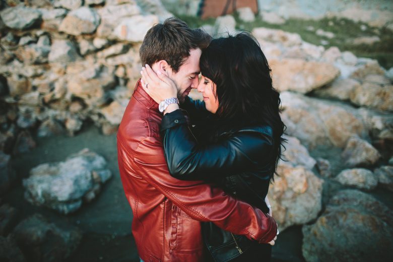 couple in leather jackets embrace with eyes closed, foreheads touching, in front of sea rocks