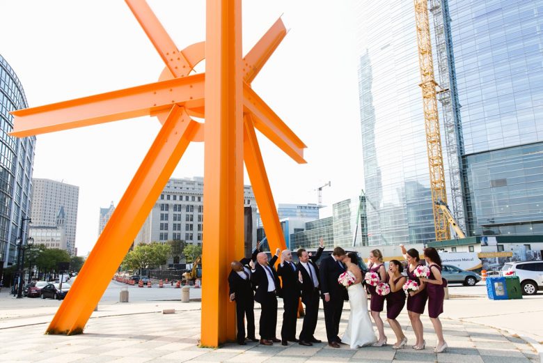 wedding party cheers on newly weds kissing beneath large orange steel urban sculpture