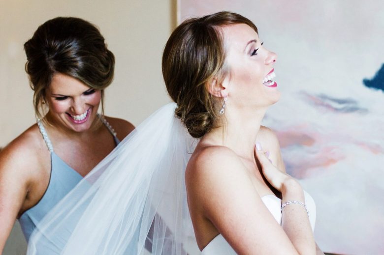 bride laughing as bridesmaid in blue dress adjusts her veil