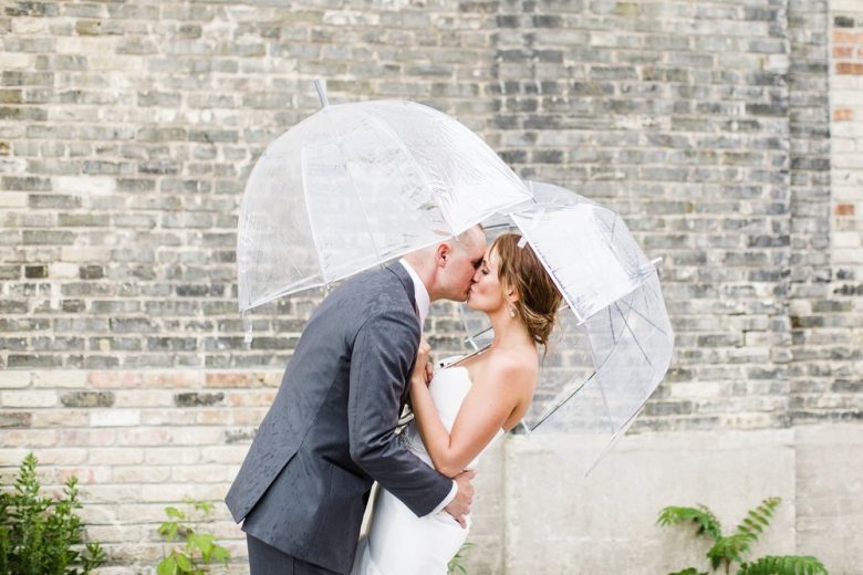 bride and groom kissing under clear plastic umbrellas in front of grey brick wall