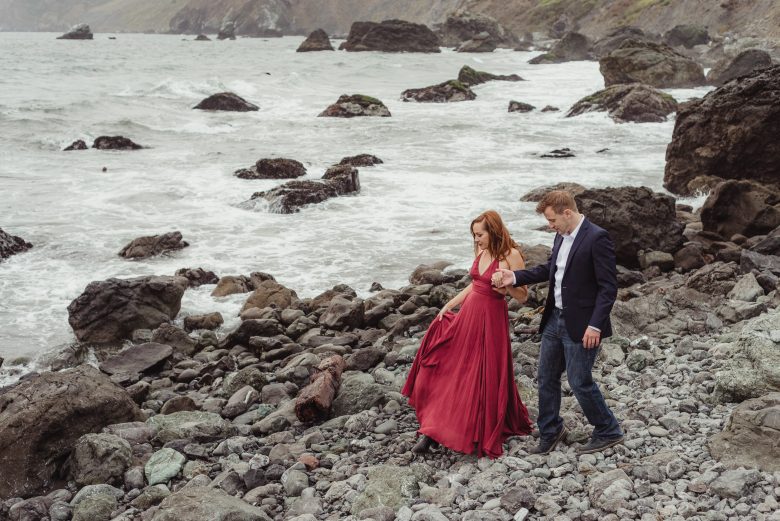 woman in red gown holds hands with man in jacket on a rocky shoreline