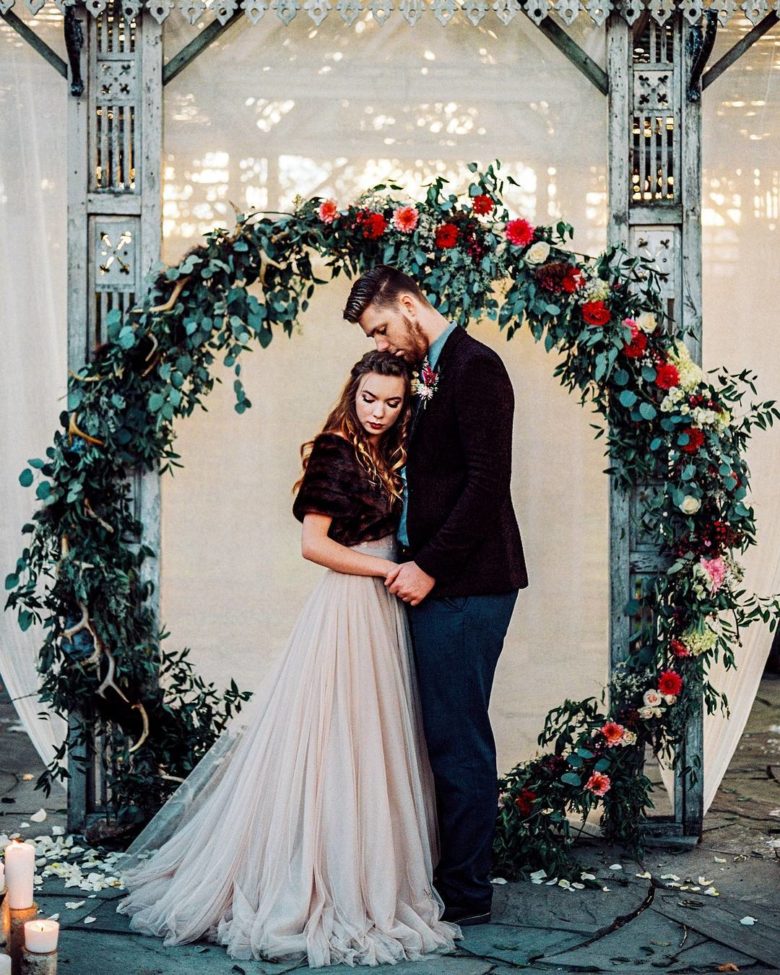 bride and groom in front of white wall, grey pillars, and large wreath of greenery, adorned on one side with red, pink, and white flowers, sitting on the ground 
