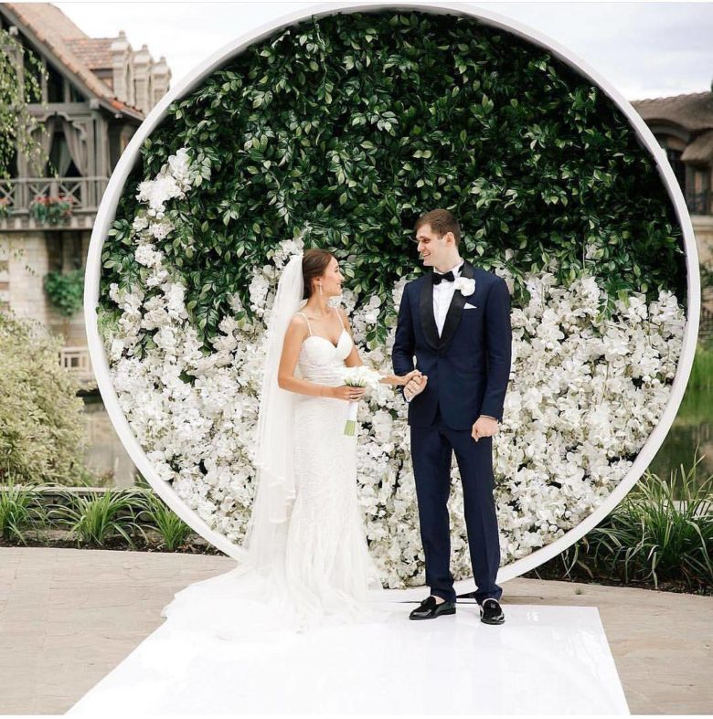 bride and groom stand in front of large circle frame filled with greenery on top and white flowers on bottom