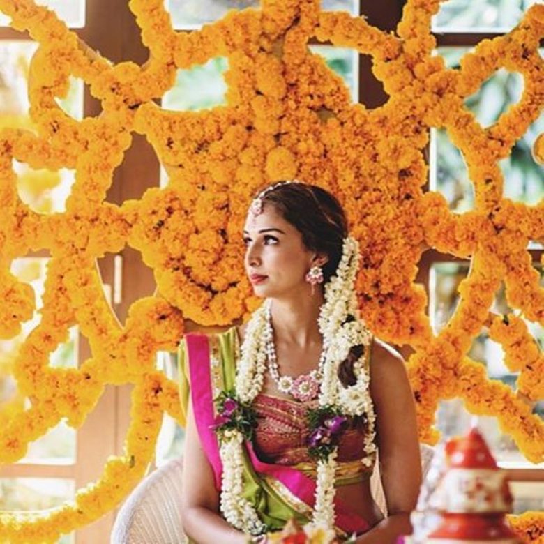 bride in bright pink and green sari sits in front of sculpturally woven wall of marigolds