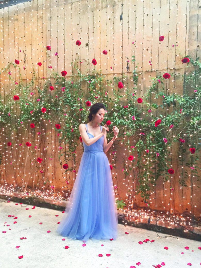 woman in ethereal periwinkle dress stands in front of a wall with green plants and vertical string lights with red flowers interspersed