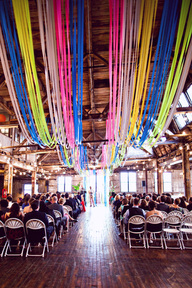 guests seated at high-ceilinged wooden wedding venue, with colorful flagging tape scalloped along ceiling