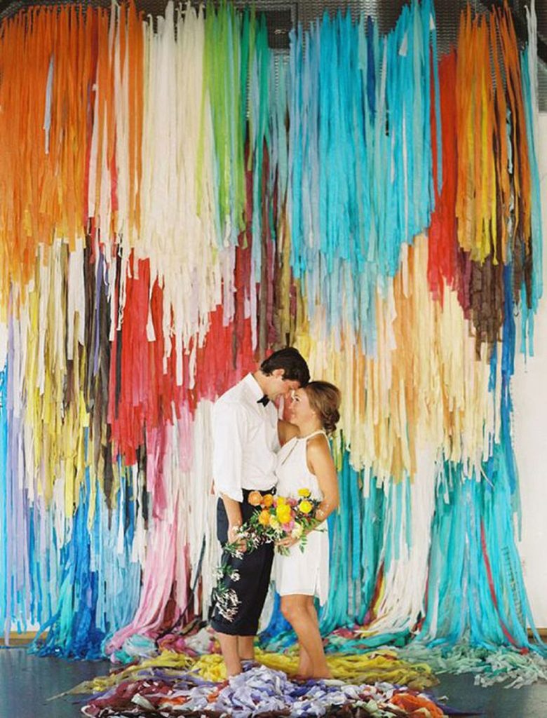 bride in short dress and groom with pants rolled up stand in front of a wall draped with lots of ribbon