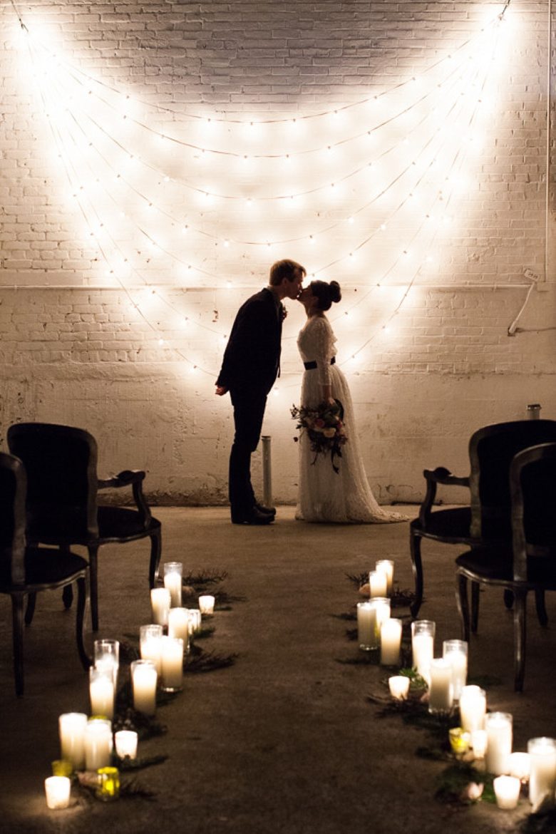 bride and groom kiss in front of a white brick wall with 7 strands of white string lights hung in a cascading scallop, aisle lined with white candles