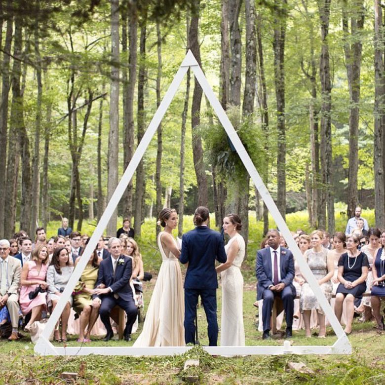 two brides with an officiant in front of guests at wedding ceremony in woods stand before a larg equilateral white triangle frame