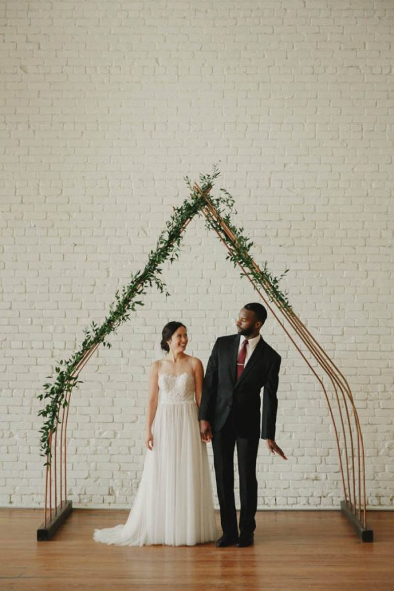 bride and groom under minimal house-shaped arch with vine greenery coming up one side and halfway down the other, in front of a white brick wall