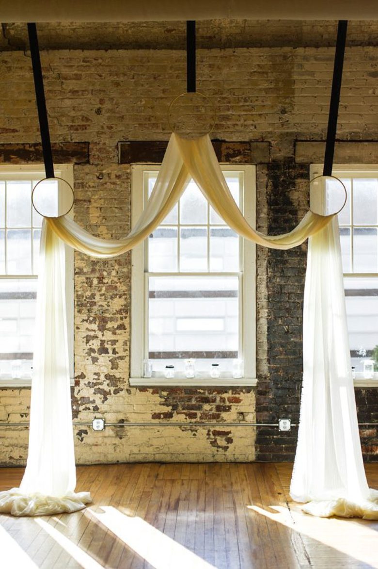 a long piece of fabric draped through three circles suspended from a loft ceiling in front of an aged brick wall with windows