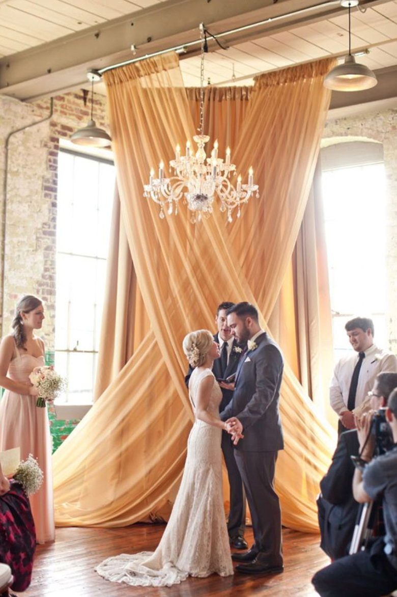 couple stands in front of officiant, underneath a chandelier in front of a large cross-section of draped fabric in a brightly lit loft 
